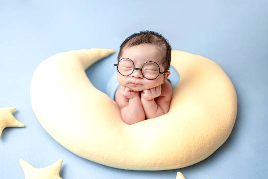 baby with eyes closed wearing huge glasses and resting on a crescent moon-shaped pillos