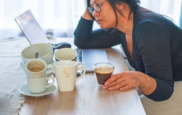 a woman looking stressed and tired drinking a cup of coffee and surrounded by multiple cups empty and full