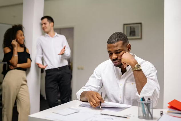 a man sitting at his desk with two co-workers talking in the background