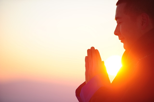 a person with eyes closed and hands in a prayer posture with sun in the background