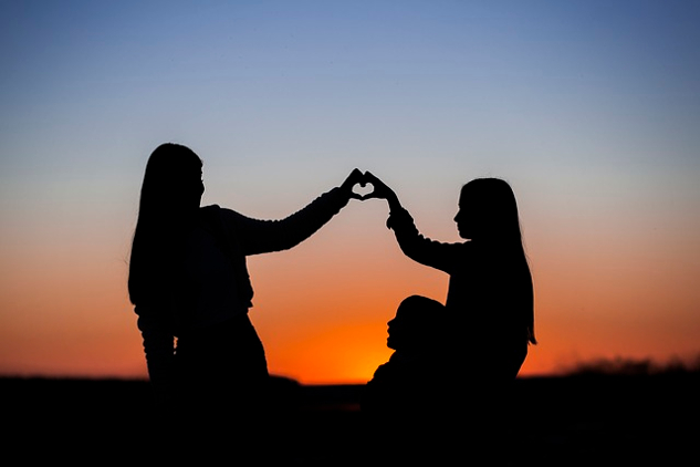 two young girls sitting in front of the sunset and making a heart with their hands