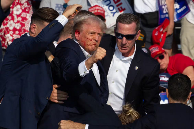 Trump with blood on his face holds up a clenched fist while people in suits surround him, illustrating the impact of social media on political violence.