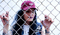 girl wearing a baseball cap standing behind a chain link fence